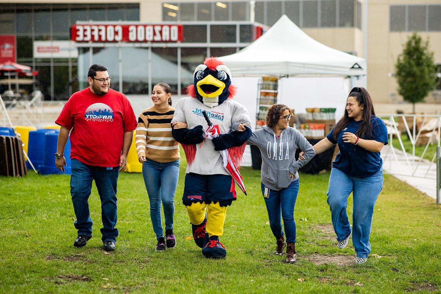 Four alumns walking with Rowdy the Roadrunner at the 同学会 Tailgate on the grass outside of the JSSB building